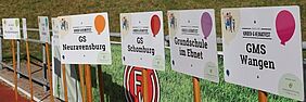 Viele Schilder mit Schulenamen, die für das Kinderfest werben. Die Schilder beinhalten Namen wie "GMS Wangen" und "GS Neuravensburg".