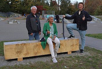 Das jüngste Projekt der Bürgerstiftung: auf dem Skateplatz wurden drei neue, einfache Sitzgelegenheiten installiert. Drei Personen stehen und sitzen an einer einfachen Sitzgelegenheit aus Holz.