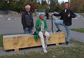 Das jüngste Projekt der Bürgerstiftung: auf dem Skateplatz wurden drei neue, einfache Sitzgelegenheiten installiert. Drei Personen stehen und sitzen an einer einfachen Sitzgelegenheit aus Holz.